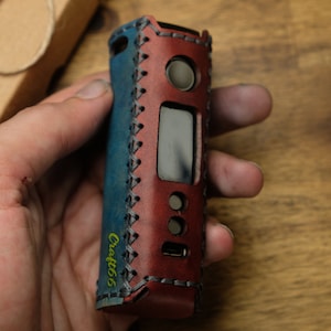 Puede incluir: Una funda de cuero azul y rojo con detalles de costura para un dispositivo electrónico de vapeo. La funda tiene una pantalla y botones. La funda está etiquetada como "Craft66".