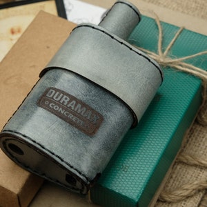 Peut inclure: Une fiole en cuir gris avec une étiquette en cuir marron qui dit "DURAMAX O CONCRETE". La fiole est posée sur une boîte verte avec un cordon marron noué autour.