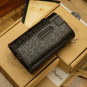 Puede incluir: Una funda de cuero negro con un patrón geométrico repetitivo y costuras visibles en los bordes. La funda presenta un recorte rectangular en la superficie frontal y está diseñada para sostener un dispositivo electrónico rectangular de forma segura.