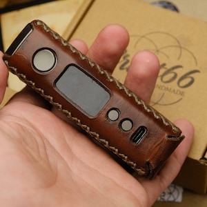 Puede incluir: Una funda de cuero marrón con detalles de costura para un dispositivo electrónico. La funda tiene una pequeña pantalla, dos botones y un puerto de carga. La funda está hecha a mano y tiene el texto "Art66 Handmade" en la caja.