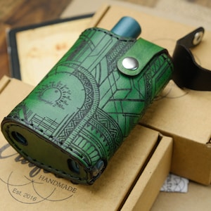 Peut inclure: Un étui en cuir vert avec un motif géométrique et une sangle en cuir noir. L'étui est doté d'une fermeture à pression en métal et est fait à la main. Le texte "HANDMADE" et "Est. 2016" est visible sur l'étui.
