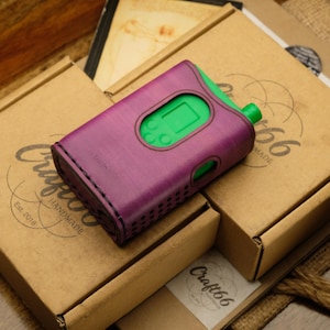 May include: A purple leather protective case with visible black stitching and a cutout for a screen and buttons, housing a bright green electronic device. The case features a perforated side panel and is resting on top of brown cardboard boxes.