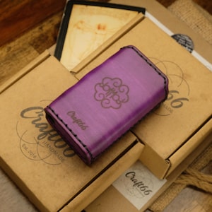 May include: A purple leather wallet with a decorative emblem and the text "Craft66" is displayed on top of several cardboard boxes. The boxes have the "Craft66 Handmade" logo and are on a wooden surface.