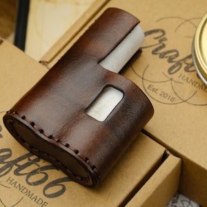 Puede incluir: Una funda de cuero marrón con costuras en los bordes. La funda tiene una abertura rectangular para un dispositivo. La funda está sobre una caja de cartón marrón con el texto "Craft66 Handmade Est. 2016" impreso en ella.