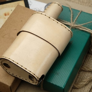 Peut inclure: Une gourde en cuir brun clair avec des détails de couture, posée sur une boîte verte avec un nœud en ficelle brune. La boîte est posée sur une boîte en papier brun avec le mot "HANDMADE" imprimé dessus.
