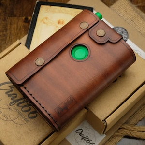 Può includere: Una custodia in pelle marrone con chiusura a patta fissata da due bottoni a pressione in metallo color bronzo. La custodia presenta un ritaglio circolare che rivela un elemento interno verde brillante. Un piccolo logo rettangolare con il testo Craft66 è impresso.