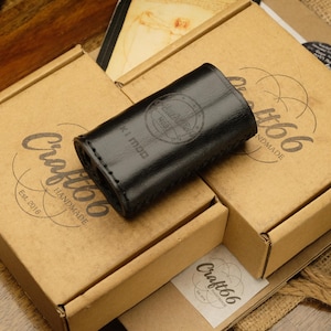 May include: A black leather case with the text "K1 Mod" and "Craft66 Handmade" is displayed on top of two cardboard boxes. The boxes have the "Craft66 Handmade" logo and the text "Est. 2016" printed on them. The background is a wooden surface.
