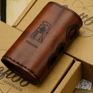 Könnte beinhalten: Ein braunes Lederetui mit einem genähten Rand und einem schwarzen Logo mit der Aufschrift "DICODES" und einer Grafik eines Gebäudes mit einem Schornstein. Das Etui liegt auf einem braunen Karton mit dem aufgedruckten Text "Craft66 Handmade Est. 2016".