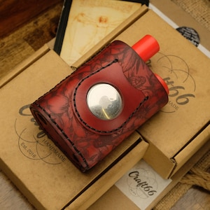 Peut inclure: Un étui en cuir rouge avec des motifs floraux et un bouton argenté, conçu pour un appareil de vapotage. L'étui a un embout buccal rouge et est posé sur une boîte en carton. La boîte porte l'inscription "Craft66 Handmade Est. 2018".