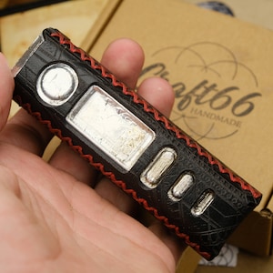 Puede incluir: Mod de vape de cuero negro con costuras rojas y detalles plateados. El mod se sostiene en una mano y tiene el texto "Craft66 Handmade" en la caja.