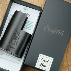 Può includere: Una custodia in pelle nera con un disegno di un edificio e il testo "DICODES" su di essa. La custodia è in una scatola nera con il testo "Craft66" e "Hand Made" su di essa.