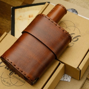 Peut inclure: Une flasque en cuir marron avec une fermeture à rabat cousue. La flasque est posée sur une boîte en carton marron avec le texte "Craft 1966 Handmade Est 2016" imprimé dessus.