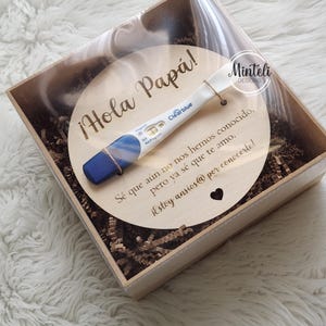 May include: A wooden box with a clear lid contains a round wooden plaque and a Clearblue pregnancy test. The plaque has the text "Hola Papá!" and a message in Spanish. The test is tied to the plaque with twine.