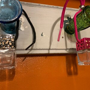 May include: Three car air fresheners hanging from a white shelf. The air fresheners are in glass bottles with metal caps. The caps are decorated with a black and white pattern, a pink and white pattern, and a green and white pattern. The air fresheners are hanging from black and pink strings.