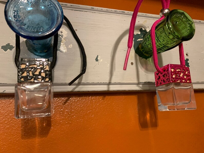May include: Three car air fresheners hanging from a white shelf. The air fresheners are in glass bottles with metal caps. The caps are decorated with a black and white pattern, a pink and white pattern, and a green and white pattern. The air fresheners are hanging from black and pink strings.