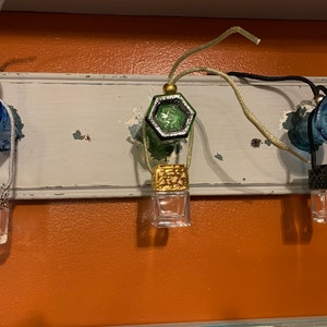 May include: Three glass car air fresheners hanging from a white shelf. The air fresheners are clear glass bottles with a decorative metal top. The tops are gold, green, and black. The shelf is attached to a wall painted orange.