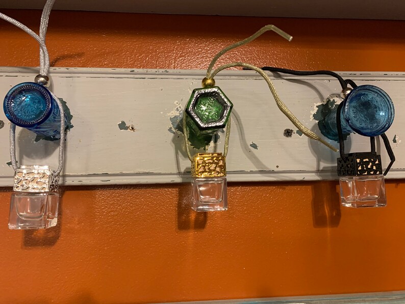 May include: Three glass car air fresheners hanging from a white shelf. The air fresheners are clear glass bottles with a decorative metal top. The tops are gold, green, and black. The shelf is attached to a wall painted orange.