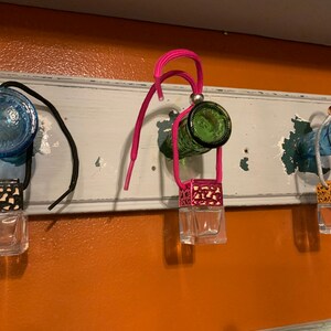 May include: Three glass bottle car air fresheners hanging from a shelf. The bottles are blue, green, and blue. Each bottle has a metal and glass diffuser attached to it. The diffusers are black, pink, and gold.