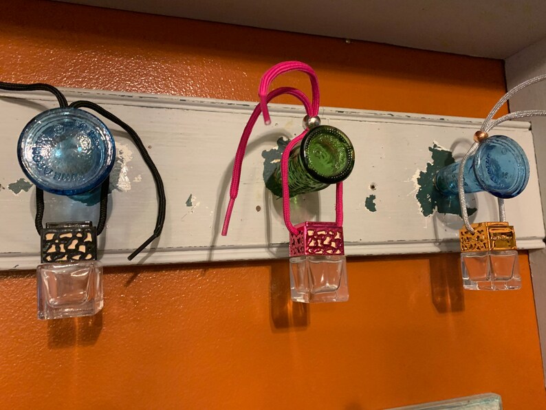 May include: Three glass bottle car air fresheners hanging from a shelf. The bottles are blue, green, and blue. Each bottle has a metal and glass diffuser attached to it. The diffusers are black, pink, and gold.