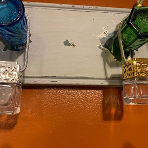 May include: Two glass car air fresheners hanging from a white wooden surface. The air fresheners are in clear glass bottles with decorative metal tops. One air freshener has a gold top and the other has a silver top.