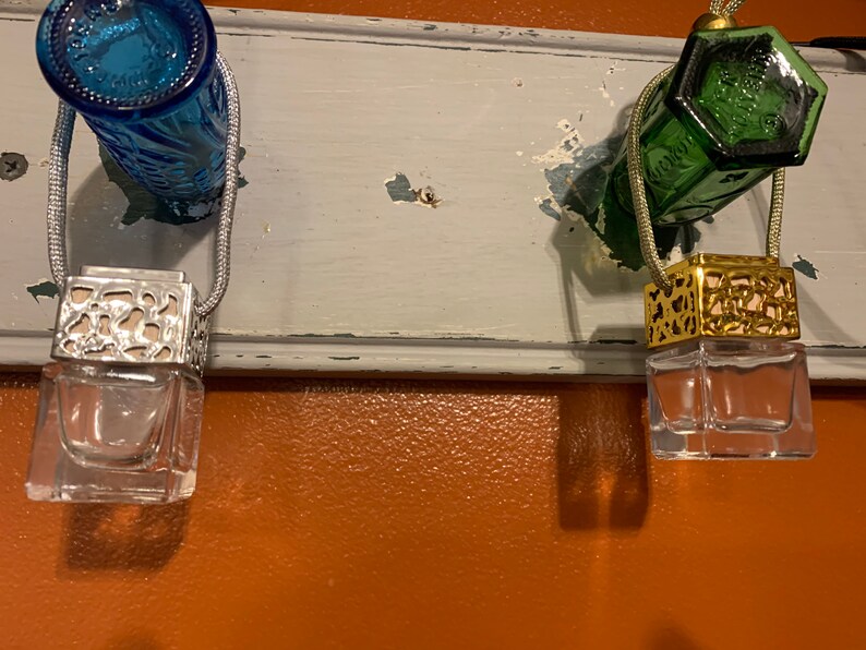 May include: Two glass car air fresheners hanging from a white wooden surface. The air fresheners are in clear glass bottles with decorative metal tops. One air freshener has a gold top and the other has a silver top.