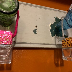 May include: Two car air fresheners hanging from a wooden surface. The air fresheners are made of glass and have a metal top. One air freshener has a pink metal top and the other has a gold metal top.