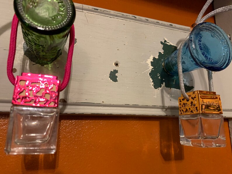 May include: Two car air fresheners hanging from a wooden surface. The air fresheners are made of glass and have a metal top. One air freshener has a pink metal top and the other has a gold metal top.