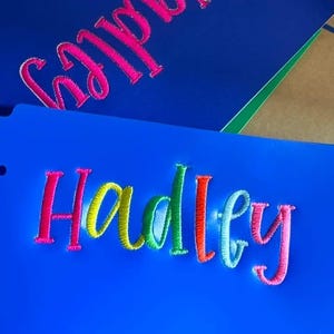 Puede incluir: Dos carpetas azules con el nombre "Hadley" bordado en hilo colorido. Una carpeta tiene el nombre en rosa, mientras que la otra tiene el nombre en un degradado de colores que incluye rosa, amarillo, verde y naranja.