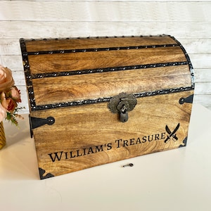 May include: A wooden treasure chest with a black metal latch and hinges. The chest is decorated with a black metal design and the words "William's Treasure" are engraved on the front. A small key is lying on the table next to the chest.