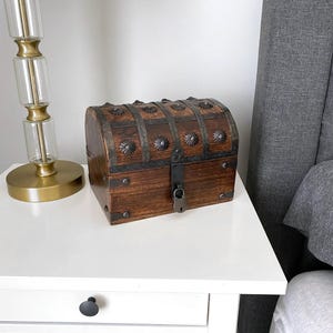 May include: A dark brown wooden chest with a curved lid, featuring decorative metal bands, circular metal studs, and a central metal latch with a small padlock. The box has a rectangular base and a hinged top, showcasing a wood grain finish.