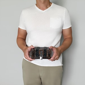 May include: A small, wooden treasure chest with metal accents and a silver padlock. The chest is rectangular with dark brown wood and black metal straps. The chest is held by a person wearing a white v-neck shirt and khaki pants.
