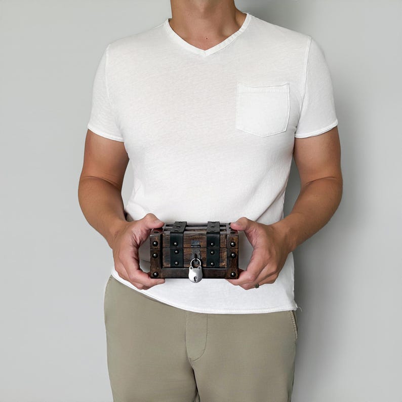 May include: A small, wooden treasure chest with metal accents and a silver padlock. The chest is rectangular with dark brown wood and black metal straps. The chest is held by a person wearing a white v-neck shirt and khaki pants.