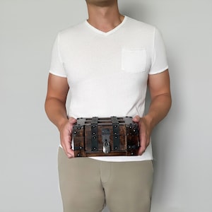 May include: A small, rectangular wooden chest with dark brown stain and metal accents. The chest has a hinged lid, metal bands, and a lock. The overall design is reminiscent of a vintage treasure chest.