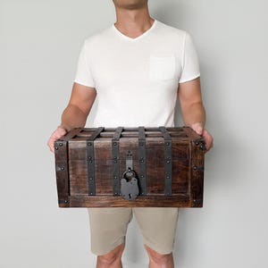 May include: A wooden chest with dark brown stain, reinforced with black metal bands and hardware. The chest features a large, antique-style lock. The chest is held by a person.