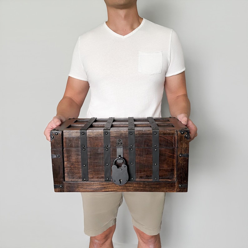 May include: A wooden chest with dark brown stain, reinforced with black metal bands and hardware. The chest features a large, antique-style lock. The chest is held by a person.