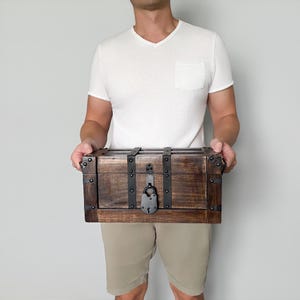 May include: A rectangular wooden chest with dark brown stain, metal accents, and a lock. The chest has metal bands and corner protectors. The chest is held by a person wearing a white v-neck shirt and khaki shorts.