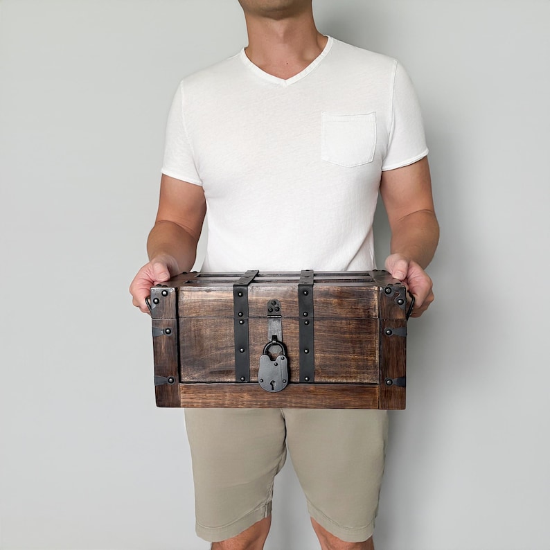 May include: A rectangular wooden chest with dark brown stain, metal accents, and a lock. The chest has metal bands and corner protectors. The chest is held by a person wearing a white v-neck shirt and khaki shorts.