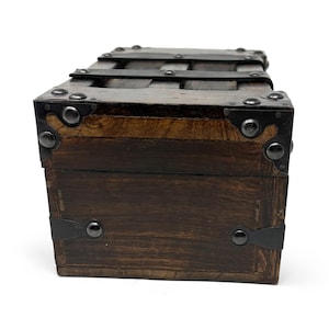 May include: A small, rectangular wooden chest with dark brown stain. The chest features metal bands and corner reinforcements with rivets. The top has a series of horizontal metal bars. The overall design suggests a vintage or antique style.