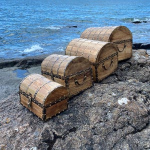Pu&ograve; includere: Quattro forzieri in legno con dettagli e cerniere in ottone, disposti in ordine decrescente di dimensioni su una superficie rocciosa. I forzieri sono in legno scuro con una finitura naturale.