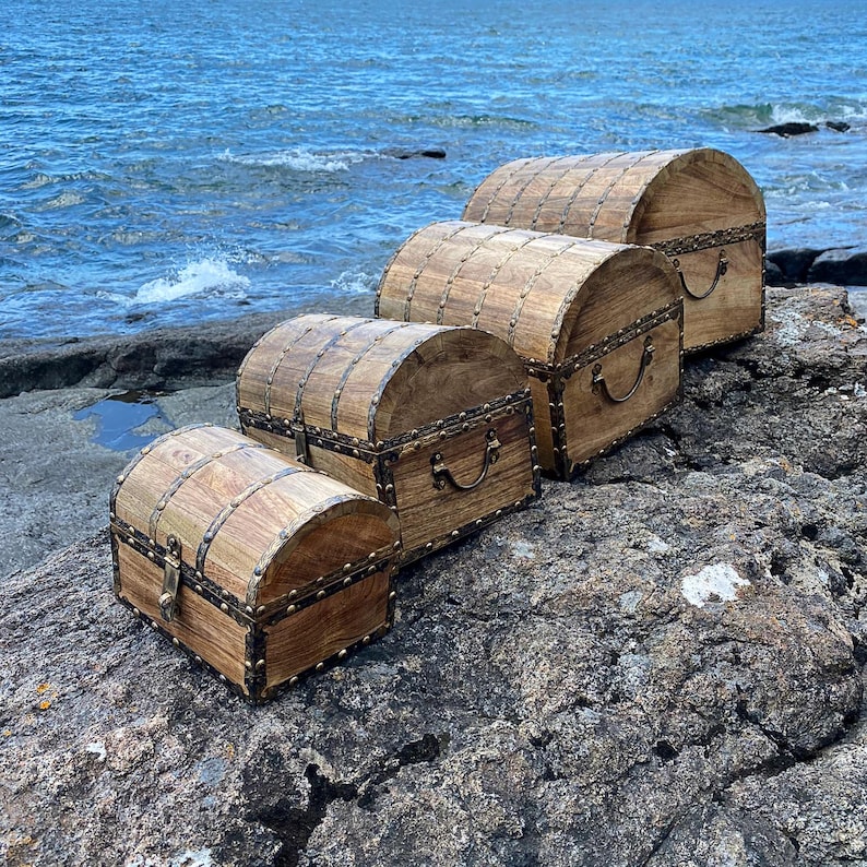 Pu&ograve; includere: Quattro forzieri in legno con dettagli e cerniere in ottone, disposti in ordine decrescente di dimensioni su una superficie rocciosa. I forzieri sono in legno scuro con una finitura naturale.