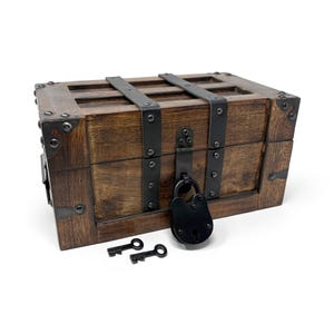 May include: A wooden treasure chest with metal accents and a black padlock. Two keys rest in front of the chest. The chest is brown with dark metal straps and corner reinforcements, suggesting a vintage or antique style.