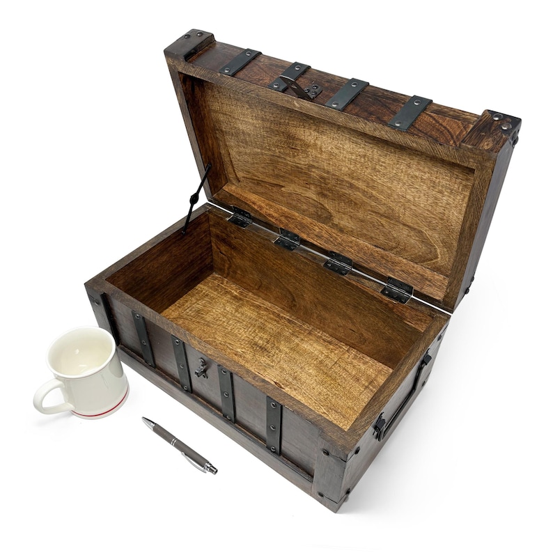 May include: An open, dark-stained wooden chest with metal accents. The chest is rectangular with a hinged lid and metal bands. A white mug with a red stripe and a pen are next to the chest.