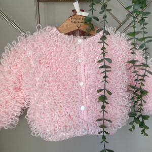 May include: A baby's cardigan in a soft pink hue, featuring a unique looped texture. The handmade garment has a round neck, long sleeves, and is fastened with three white buttons. It is displayed on a wooden hanger.