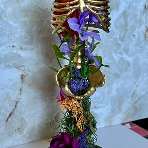 May include: A gold-colored, decorative human torso sculpture with a red interior, adorned with purple and green artificial flowers and moss. The sculpture is mounted on a black, round base covered in green moss.