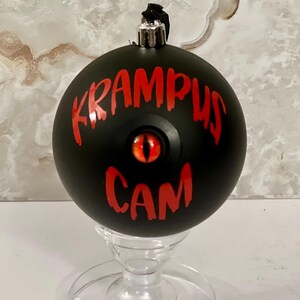 May include: A matte black spherical ornament featuring the red text KRAMPUS CAM and a central red eye with a vertical black pupil. The ornament has a silver metal cap at the top with a black hanging loop and rests on a clear glass stand.
