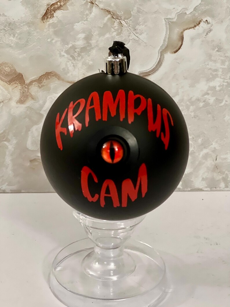 May include: A matte black spherical ornament featuring the red text KRAMPUS CAM and a central red eye with a vertical black pupil. The ornament has a silver metal cap at the top with a black hanging loop and rests on a clear glass stand.