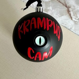 May include: A matte black spherical Christmas ornament featuring the red text KRAMPUS CAM and a central, realistic green eye with a vertical black pupil. The ornament has a silver metal cap and a black hanging loop at the top for display.