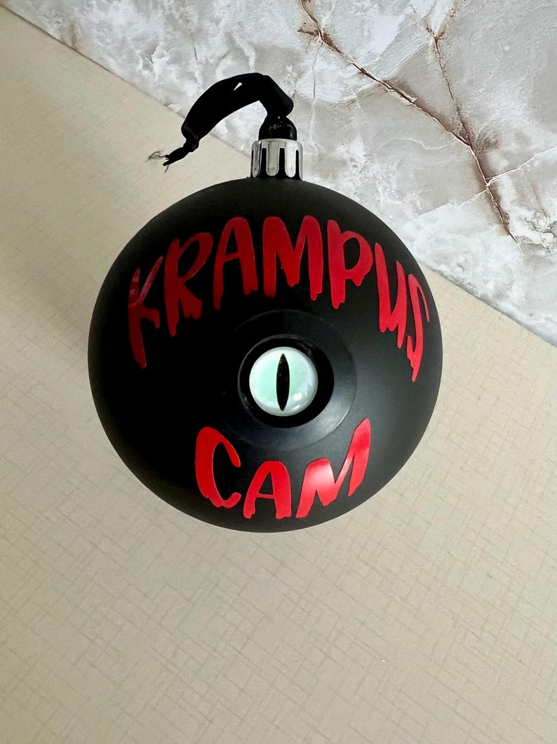 May include: A matte black spherical Christmas ornament featuring the red text KRAMPUS CAM and a central, realistic green eye with a vertical black pupil. The ornament has a silver metal cap and a black hanging loop at the top for display.