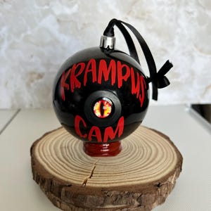 May include: A spherical black ornament featuring the red text KRAMPUS CAM and a central eye graphic with a yellow iris and black pupil. The ornament has a silver cap, a black ribbon loop, and is supported by a small red base.