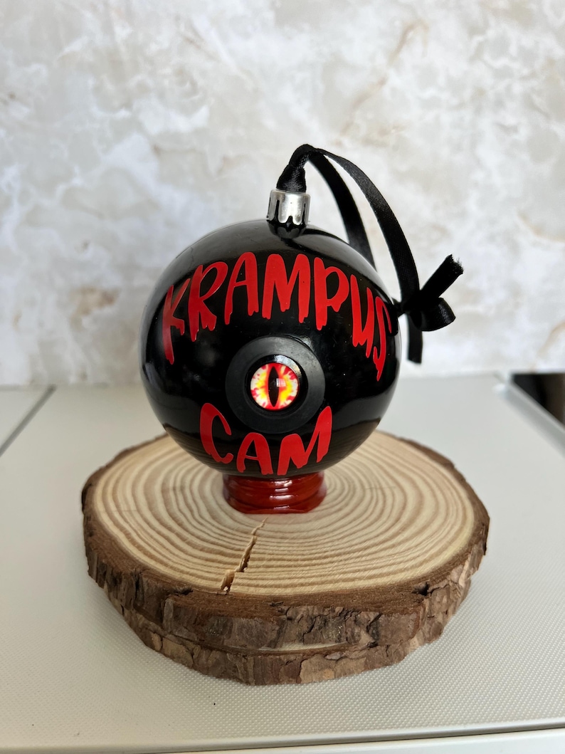 May include: A spherical black ornament featuring the red text KRAMPUS CAM and a central eye graphic with a yellow iris and black pupil. The ornament has a silver cap, a black ribbon loop, and is supported by a small red base.